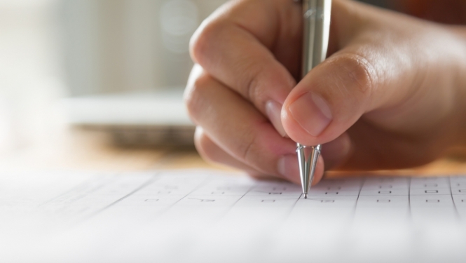 Cilvēka roka tur pildspalvu un atzīmē izvēlēto atbildes variantu lapā - Ilustratīvs attēls.