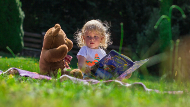 Ilustratīvs attēls - bērns sēž pļavā ar lācīša rotaļlietu 